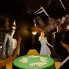 A group of Zois in InZOI playing poker and using a money machine to fire money around a dimly lit room. They have party hats on.