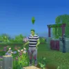 A green plantsim in The Sims 4 tending to plants in naturre. He looks to be talking to his crops while gardening in Henford on Baggley. He has a blue stripey tshirt and greens. In the background is a cottage white a stone and wood gate, plus trees and a blue sky.