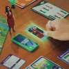 A close-up of Goliath Game's The Sims Board Game showing a hand grab a card while Bella Goth looks at them.