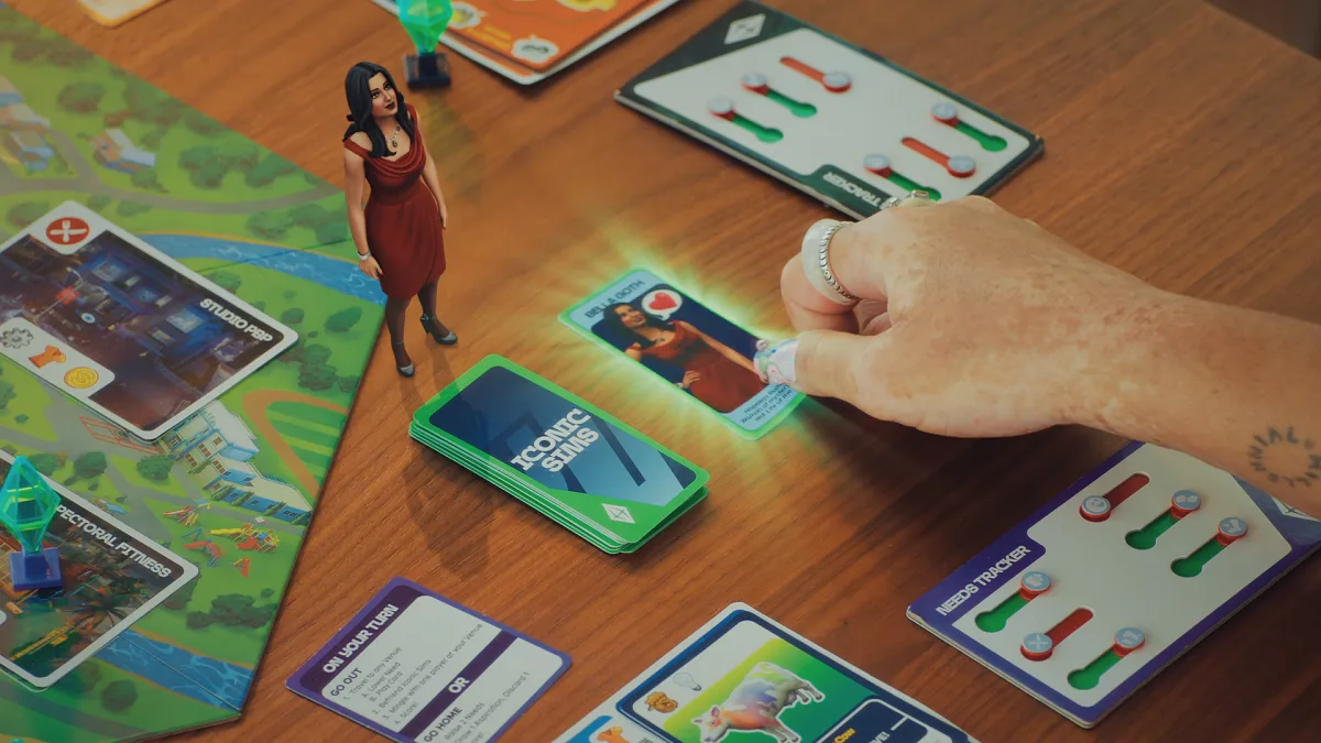 A close-up of Goliath Game's The Sims Board Game showing a hand grab a card while Bella Goth looks at them.