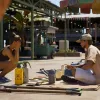 An image of a tropical street, a man is sitting cross-legged selling items and a woman is squatting across from him to enquire.