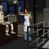 A woman in a lab coat stands in front of a robot who's sat up in surprise as if they've just been activated.