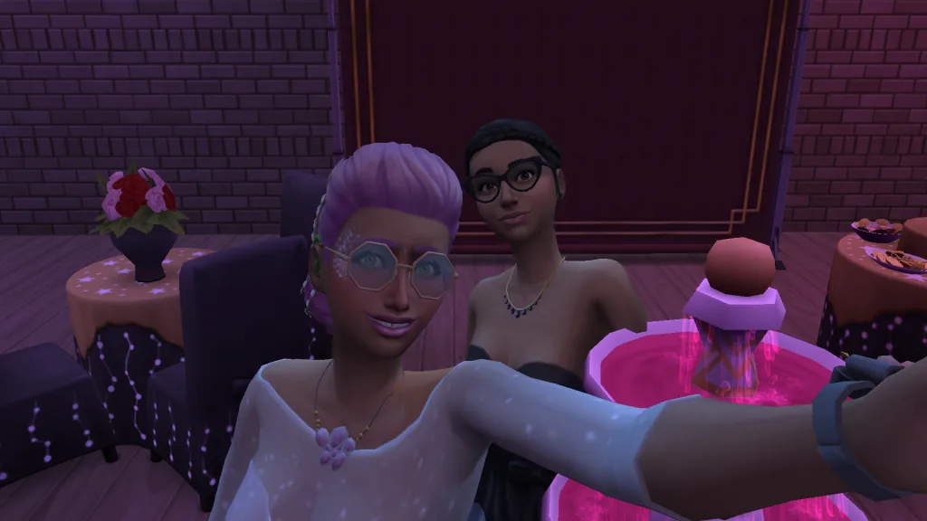 Two Sims, including Cassandra Goth, taking a selfie at prom by the punchbowl. The Sim in the front is wearing all pink while Cassandra is dressed in black.