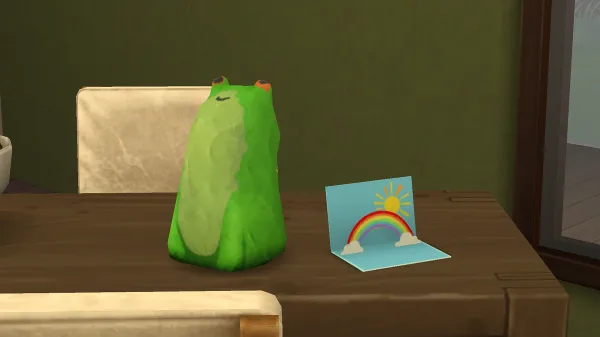 A papier-mache frog on a table next to a pop-up rainbow celebration card. in The Sims 4: Adventure Awaits.